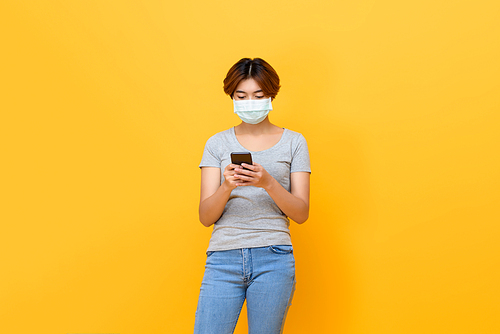 Young Asian woman wearing medical face mask using mobile phone isolated on yellow studio background