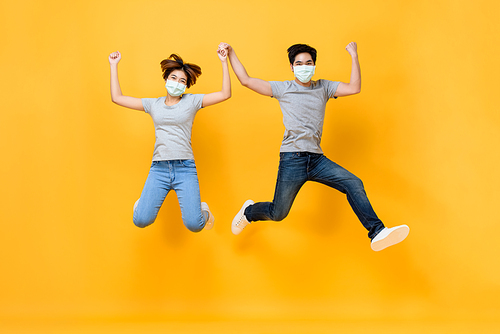 Young Asian couple wearing medical face masks and jumping with arms raised isolated on yellow background, Coronavirus protection concept