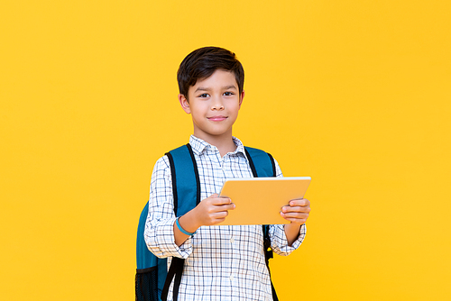 Waist up portrait of handsome young schoolboy wearing backpack holding a tablet while looking straight in camera in yellow isolated studio background