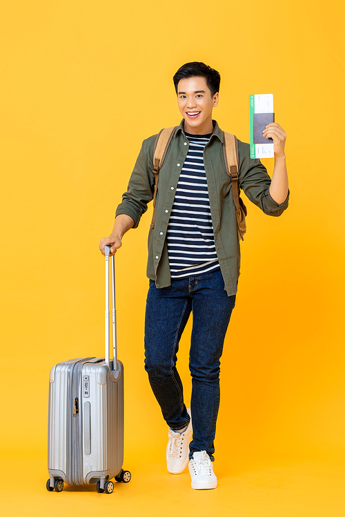 Young handsome Asian tourist man with luggage passport and boarding pass ready to go for travel on isolated yellow background