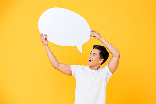 Portrait of cheerful smiling young handsome Asian man holding and looking up at speech bubble with empty space for text on colorful yellow studio background