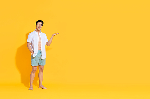 Young smiling handsome Asian man in beach attire doing open palm gesture showing empty space  in summer yellow isolated studio background