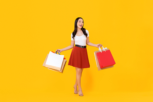 Full length portrait of young beautiful Asian woman holding colorful shopping bags in yellow isolated studio background