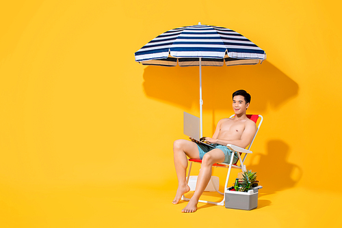 Shirtless Asian man working  on laptop computer while sitting on beach chair with umbrella isolated on yellow backgound with copy space for summer vacation concept