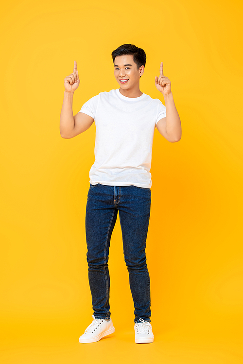 Smiling young handsome Asian man standing and pointing two fingers up isolated on yellow studio background