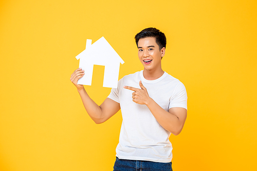 Handsome Asian man holding and pointing to house cutout isolated on yellow studio background