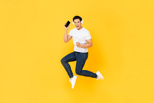 Happy young Asian man wearing headphone listening to music from mobile phone and jumping isolated on yellow background