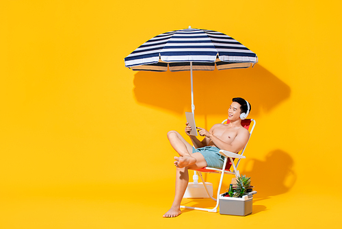 Portrait of young shirtless Asian man sitting on beach chair relaxing and listening to music in isolated summer yellow background