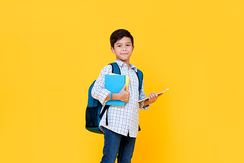 Handsome 10 year-old boy with backpack holding books and tablet computer in yellow background for education concept