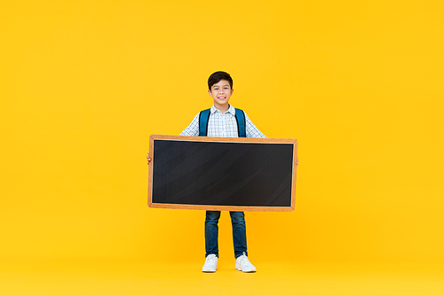 Smiling handsome 10 year-old mixed race boy holding empty blackboard isolated on yellow studio background for education concept