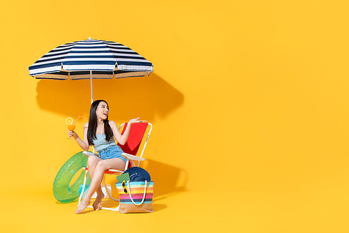 Surprised beautiful young Asian woman sitting on beach chair doing an open palm gesture isolated on bright yellow studio background with copy space
