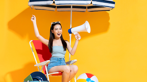 Ecstatic young Asian woman in summer beach outfit holding and announcing on megaphone in sunny yellow isolated studio background