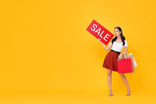 Beautiful Asian woman with shopping bags showing red sale sign isolated on yellow background with copy space