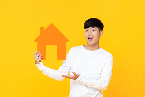 Smiling handsome Asian man holding house sign with hand presenting gesture isolated on yellow background