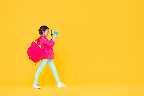 Smiling cute pretty girl with pigtail hair in pink jacket and backpack looking through a magnifying lens in yellow isolated studio background