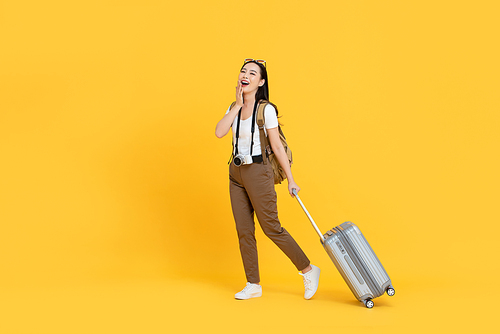 Happy young Asian tourist woman with baggage going to travel on holidays isolated on yellow background