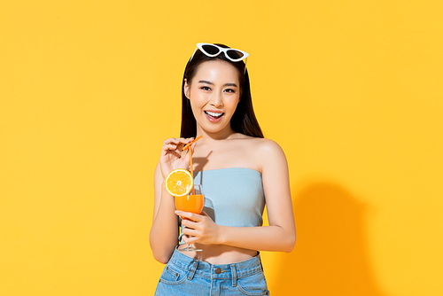 Beautiful smiling Asian woman in summer outfit with a glass of orange juice isolated on colorful yellow background