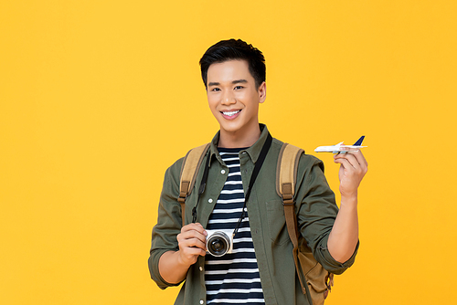 Young handsome smiling Asian tourist man holding plane model and camera isolated on yellow background
