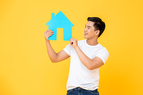 Handsome Asian man holding and looking at house cutout isolated on yellow studio background