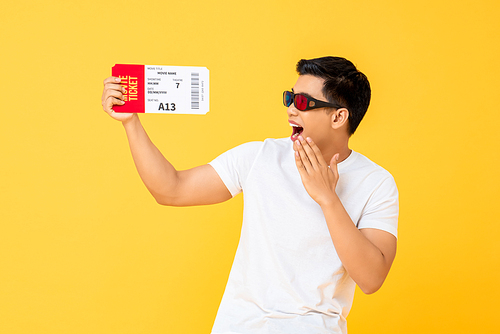 Fun portrait of surprised young Asian man wearing 3D glasses holding movie ticket in isolated studio yellow background