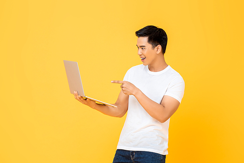Portrait of a smiling  young handsome Asian man holding laptop computer while looking and pointing on screen in isolated studio yellow background