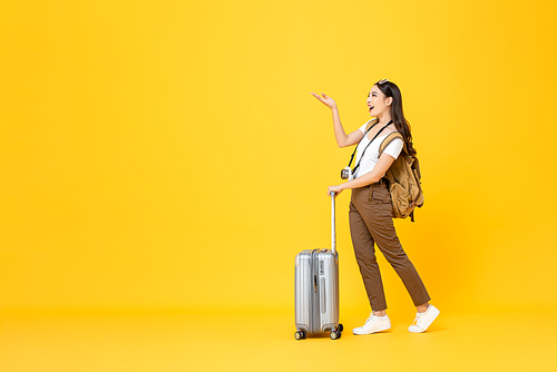 Full length portrait of beautiful smiling young Asian woman tourist with trolley bag looking and opening hand to copy space isolated on colorful yellow studio background