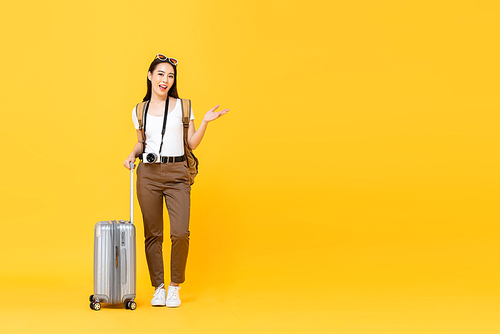 Full length portrait of smiling cute young Asian woman tourist with trolley bag opening hand to copy space aside isolated on yellow studio background
