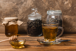 Cup of tea near honey and blurred jars on wooden surface on textured stone background