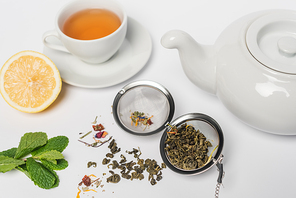Dry tea in infuser near mint, lemon and cup on white background