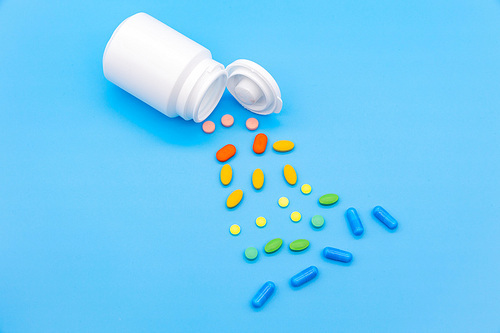 파란 배경지 위에 흰색 약병이 엎어져있고 알록달록 약들이 바닥에 엎질러진 모습의 탑뷰 사진