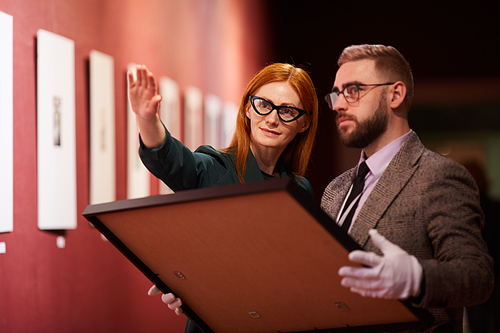 Young red haired art manager showing the place for new painting to her colleague in the gallery
