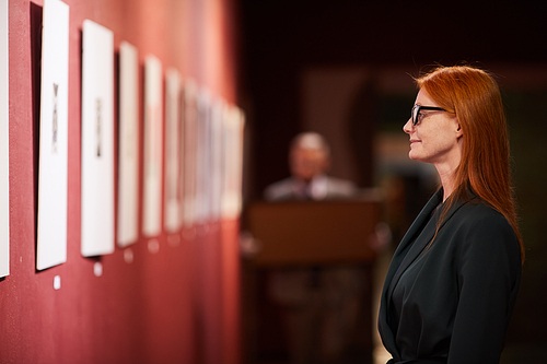 Side view of young red haired woman in eyeglasses standing in front of the wall with paintings and enjoying the art