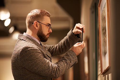 Side view of young bearded man in eyeglasses taking photo of paintings on his mobile phone in the gallery