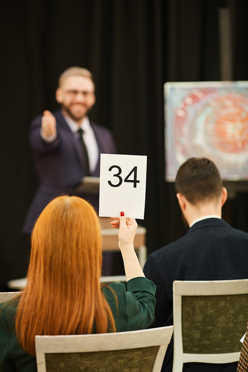 Rear view of red haired woman sitting and raising her sign and man pointing at her during auction