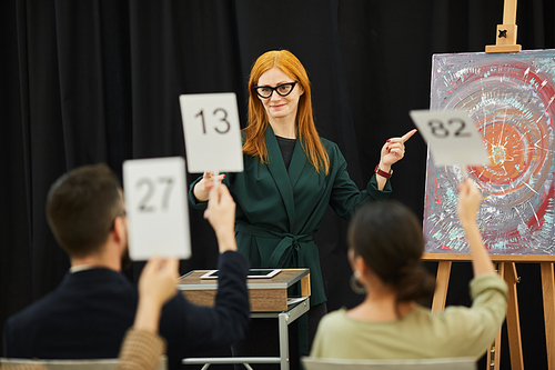 Young beautiful woman in eyeglasses presenting the painting to the audience and they raising their signs with numbers at auction