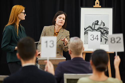 Woman standing near the painting and pointing at man who raising his sign during auction