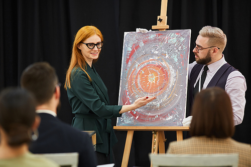 Businessman and businesswoman pointing at painting and presenting new modern art to other people at conference