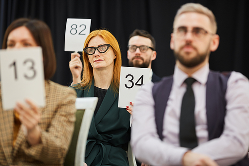 Red haired woman in eyeglasses raising sign with number while sitting among other people at auction