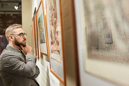 Rear view of young pensive man in eyeglasses standing and looking at paintings on the wall at gallery