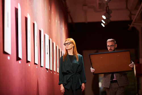 Young woman walking along the corridor with paintings on the wall together with her colleague who carrying the picture