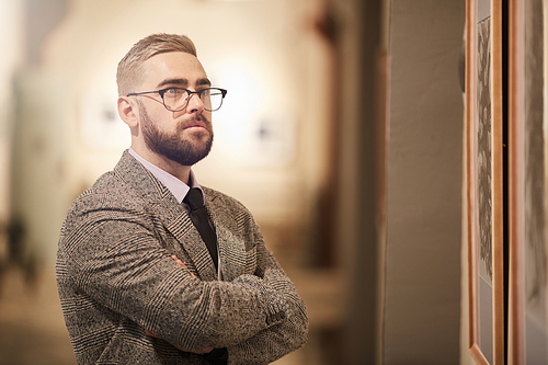 Young bearded man in eyeglasses standing with his arms crossed and looking at beautiful picture