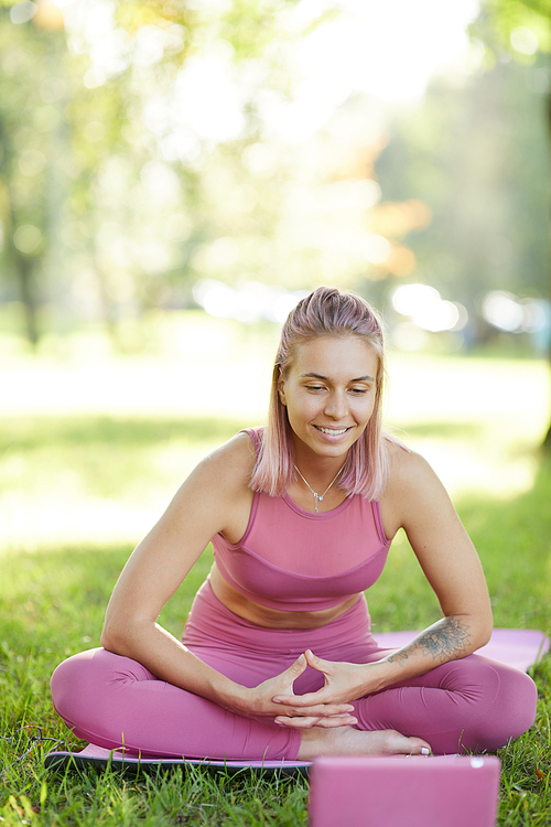 Young beautiful fit woman sitting on the grass on exercise mat looking at tablet pc and smiling she holding online training outdoors