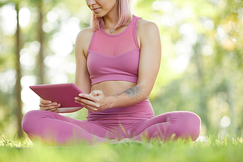 Close-up of slim sporty woman in pink sports clothing sitting on the green grass and using digital tablet during sports training outdoors