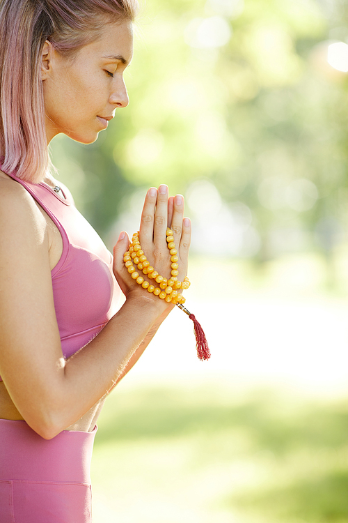 Side view of young healthy woman with amulet in her hands standing with eyes closed and meditating during yoga outdoors