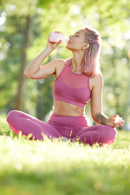 Young healthy woman in pink sports clothing sitting on the green grass and drinking water after sports training