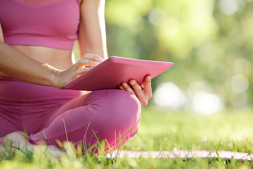 Close-up of young girl in pink clothing sitting on the green grass and using digital tablet during her sports training outdoors
