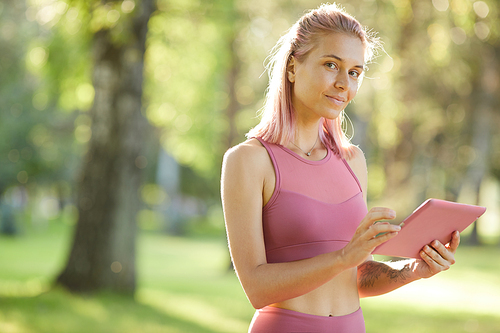 Portrait of young blonde girl looking at camera while communicating online on her tablet pc in the park
