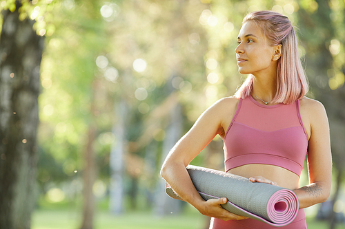 Young healthy girl in pink top holding exercise mat and enjoying the good weather in the forest