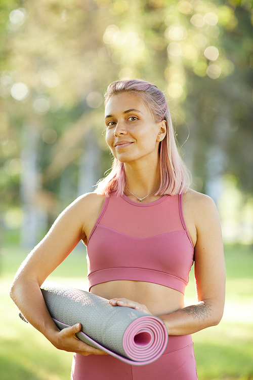 Portrait of beautiful healthy woman in pink top holding exercise mat and smiling at camera while standing outdoors in the park