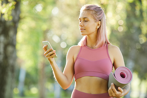 Young blonde girl in pink top holding exercise mat and typing a message on her mobile phone while standing outdoors in the forest